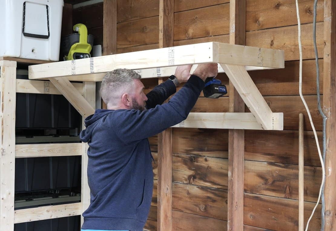 Driving screws through the 2x4 ledger into the wall studs to hang the garage shelf