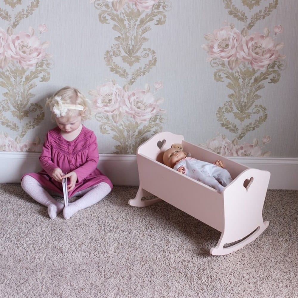 Little girl reading next to a pink painted wooden doll cradle with heart cutouts and a baby doll inside